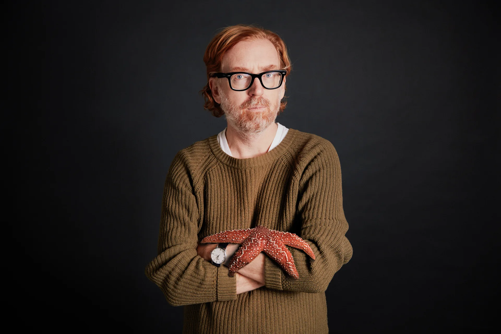 Portrait of a man in glasses and green sweater holding a red starfish across his arms